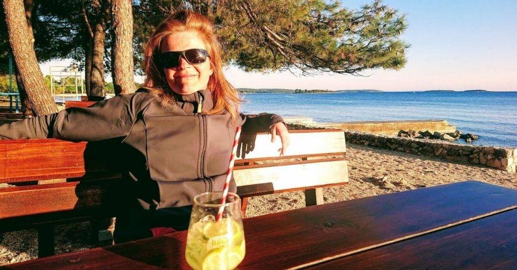 Frau auf Bank sitzend mit Sonne im Gesicht und Drink vor sich